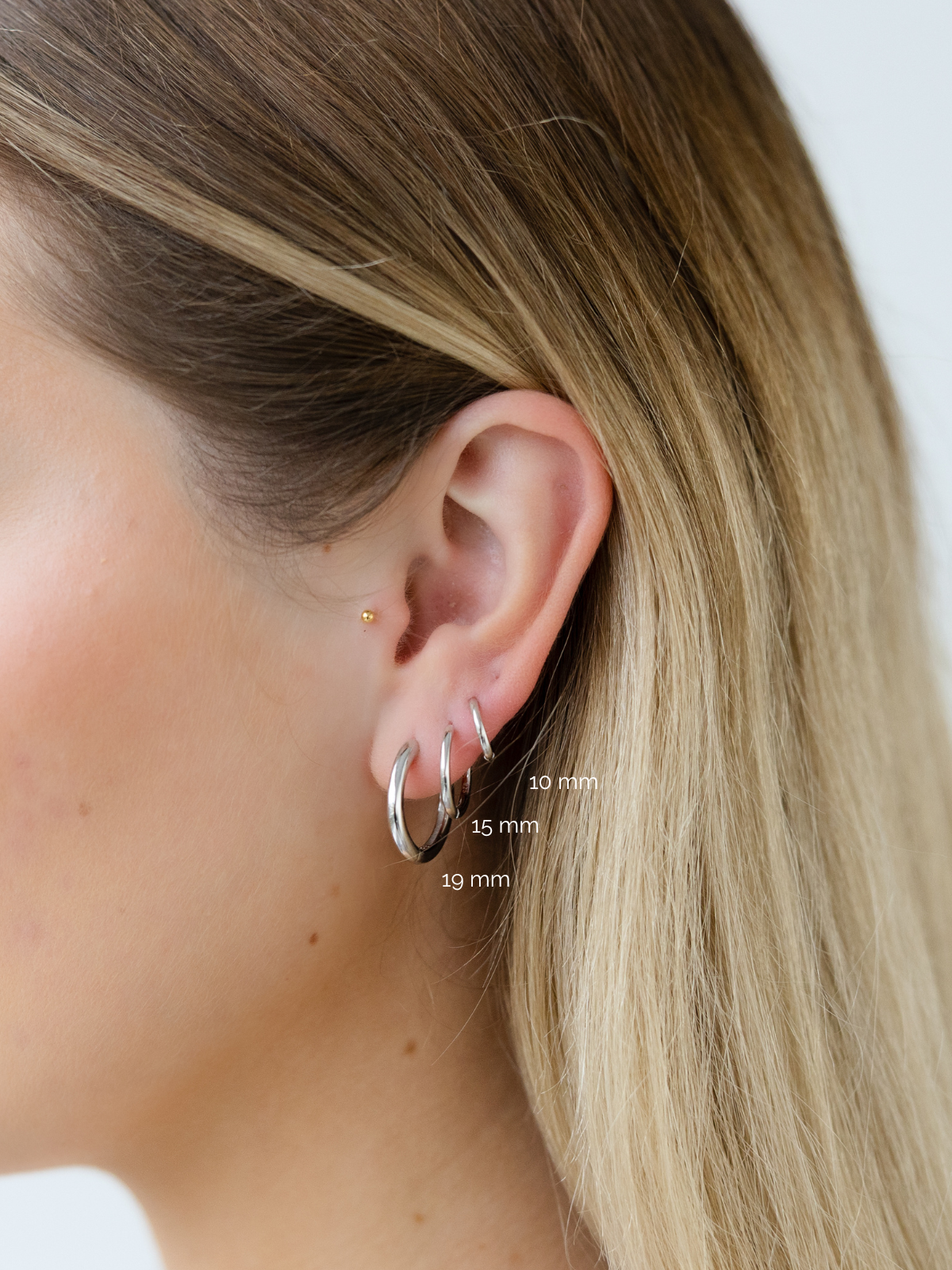 Close-up of an ear wearing a silver hoop earring with size labels on a plain background