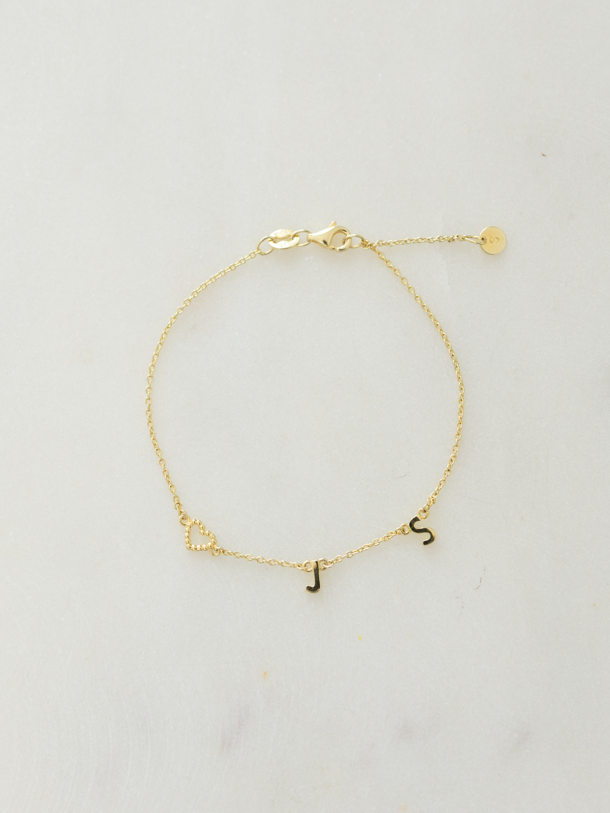 Gold choose your letters bracelet with the initials J, S & a dotty love heart pendant on a light gray background