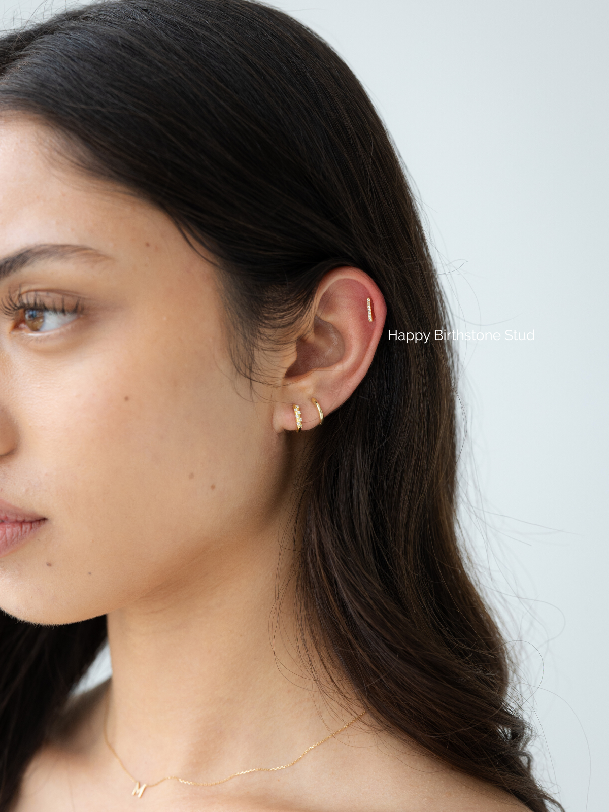 Close-up of a woman wearing a gold stack of earrings with a plain background. Features the happy birthstone earring