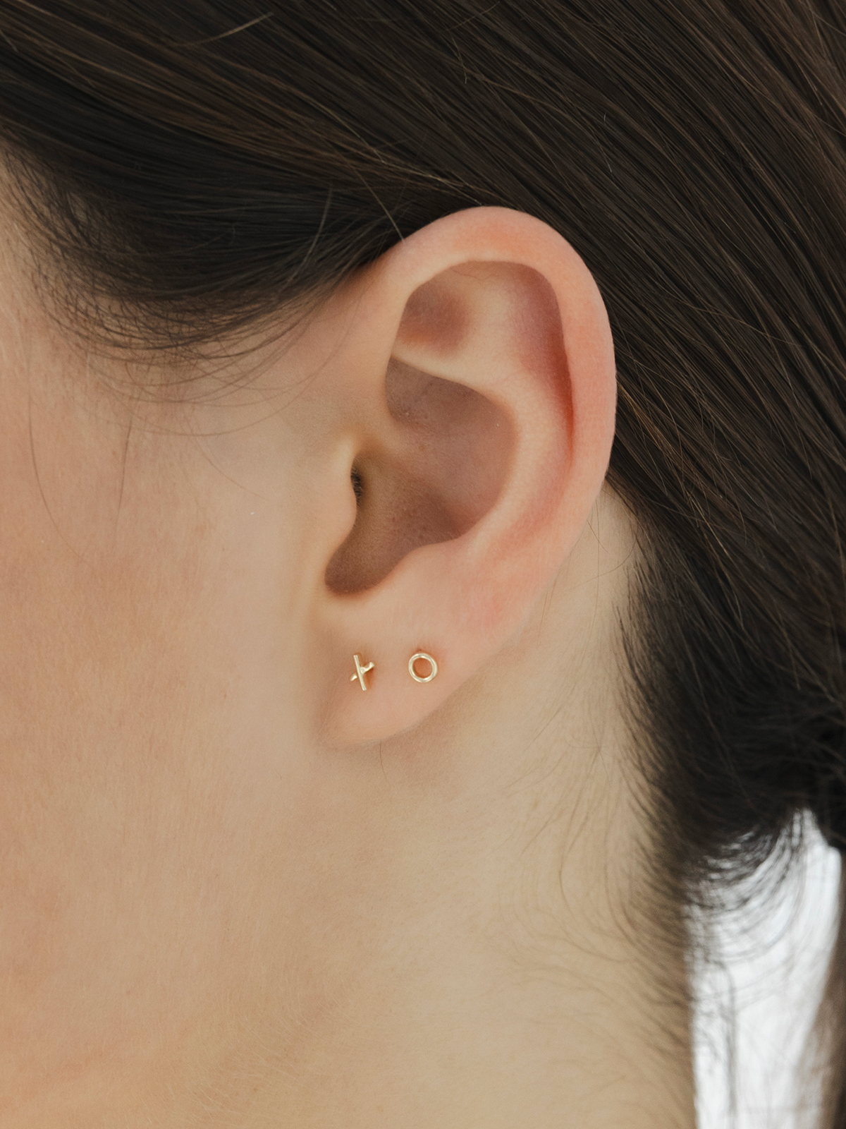 Close-up of an ear wearing solid gold 'X' and 'O' stud earrings with dark hair.