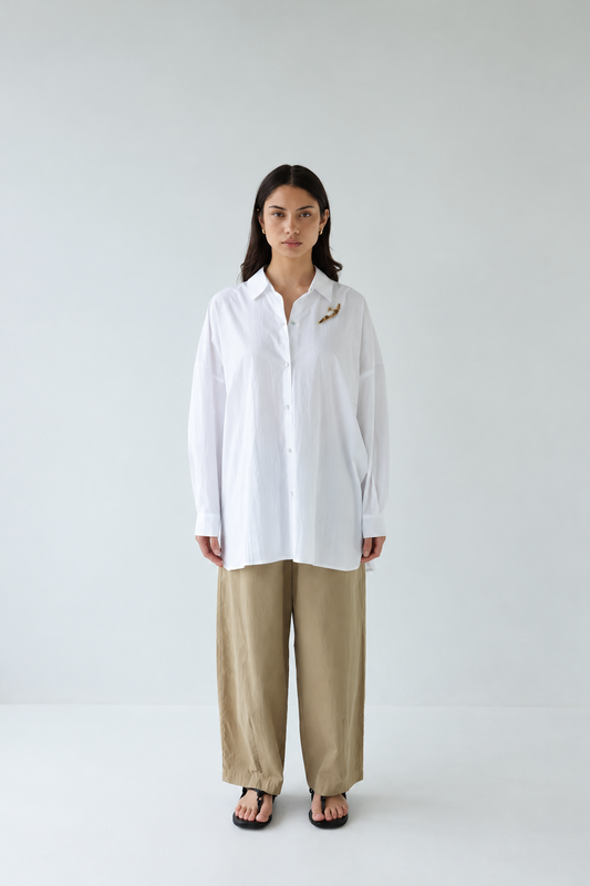 A woman stands against a plain light gray studio background wearing a loose white button-up shirt with a small gold embroidered detail on the chest, beige wide-leg pants, and black sandals.
