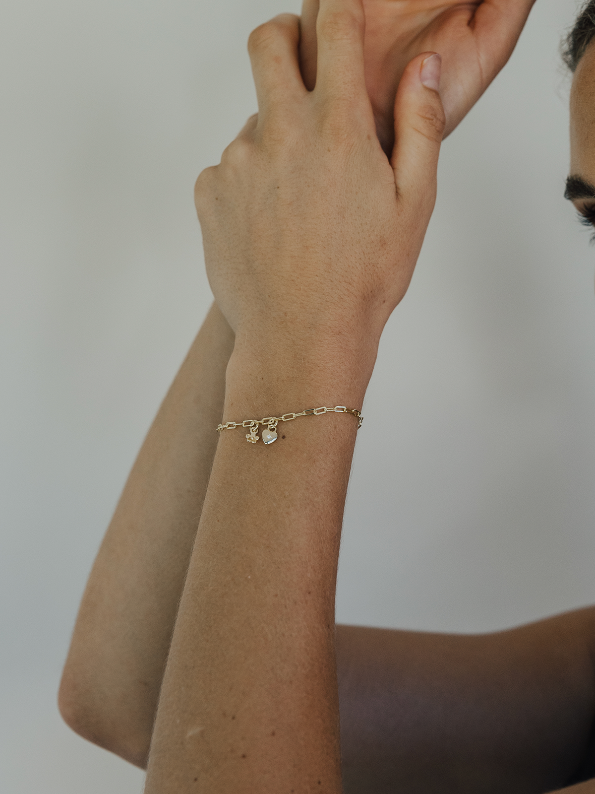 Close-up of a person wearing a gold bracelet with a sweetheart & daisy day charm on a neutral background
