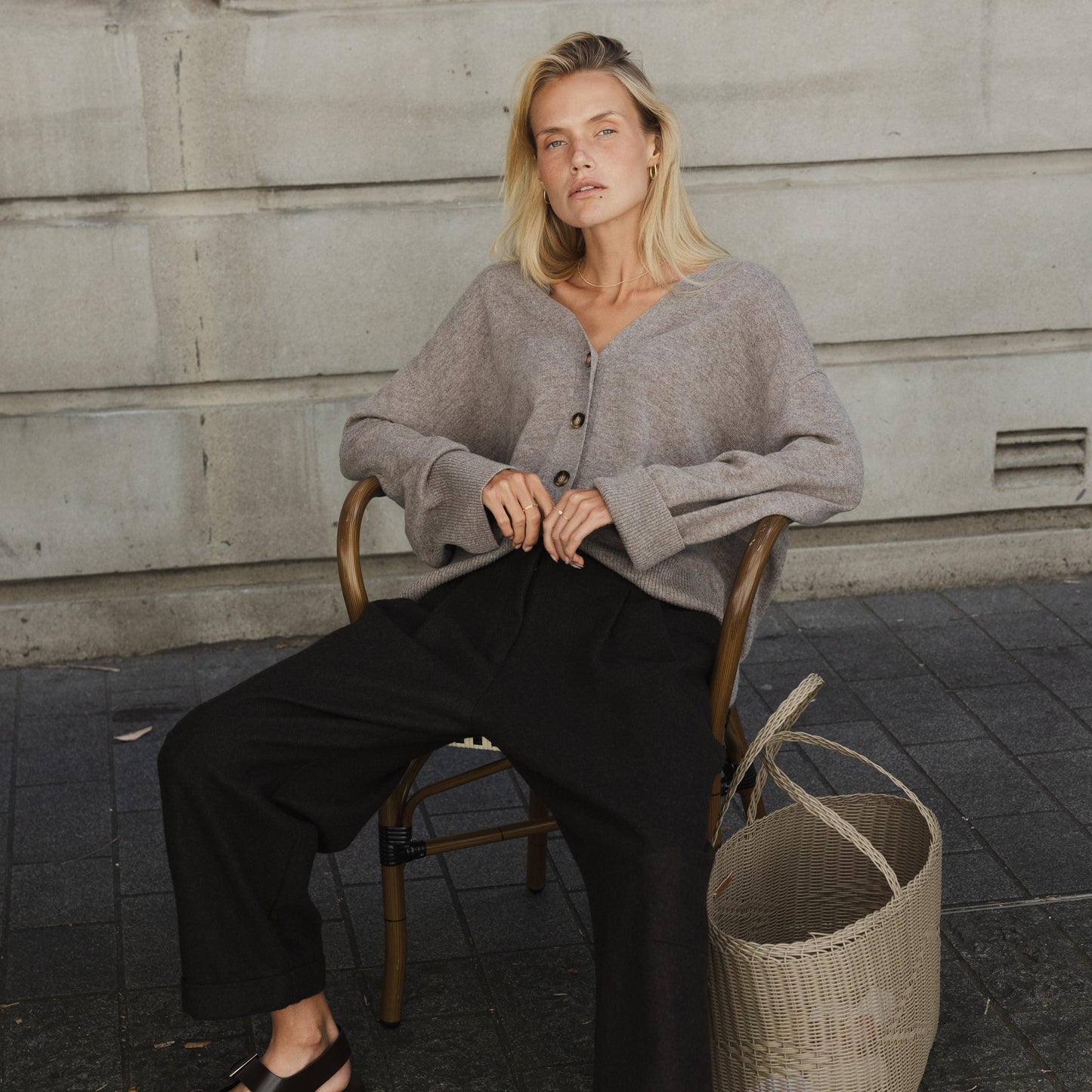 A woman seated wearing black pleated pants with a cuff and double pleats, paired with a grey knit sweater. She is accessorized with a belt and is seated next to a woven basket.