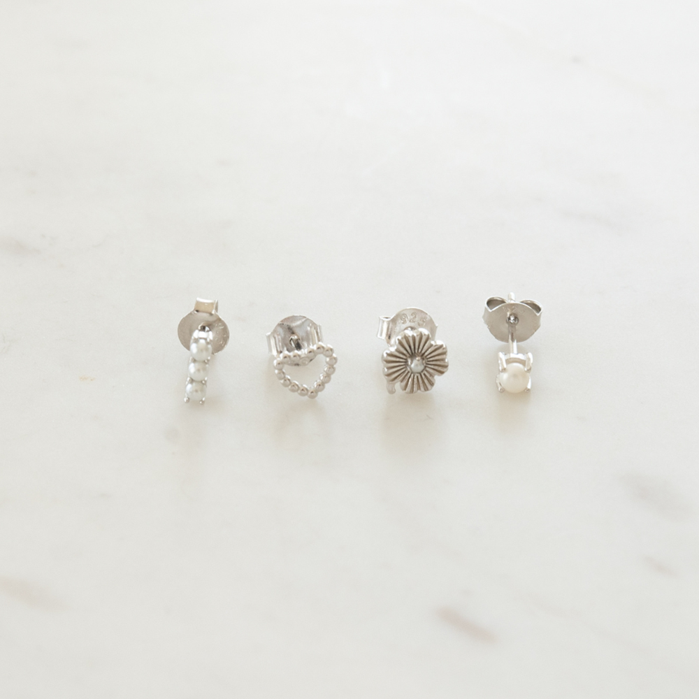 A set of four silver stud earrings with different designs including a heart, a pearl, a floral pattern, and a barbell style on a marble surface.