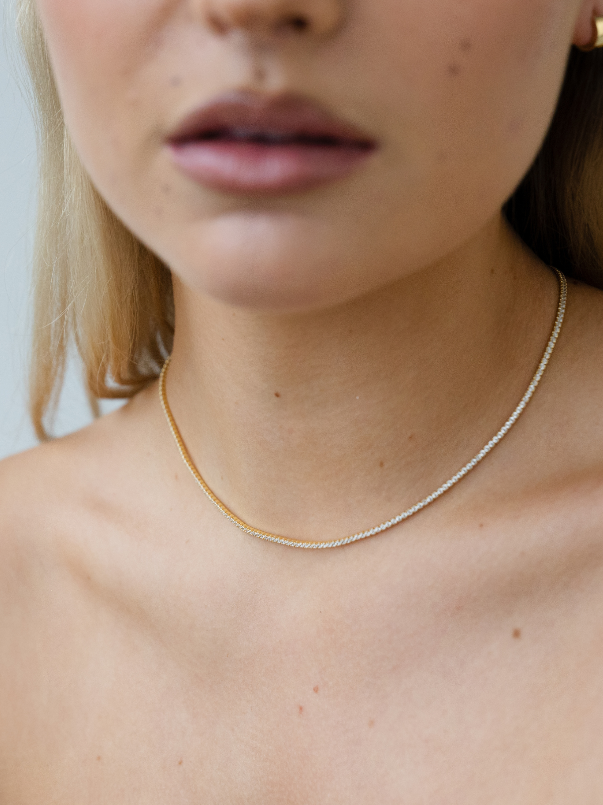 Close-up of a person wearing a delicate gold tennis chain necklace against a neutral background