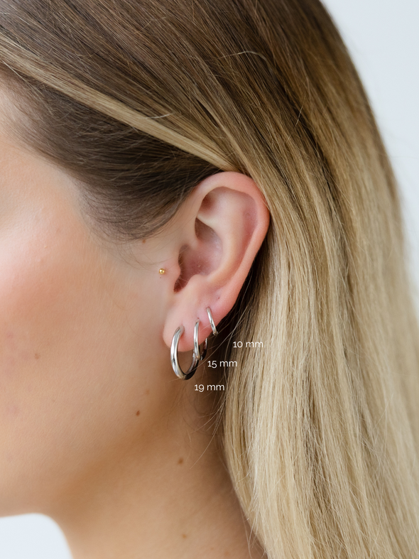 Close-up of an ear wearing a silver hoop earring with size labels on a plain background