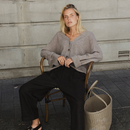 A woman seated wearing black pleated pants with a cuff and double pleats, paired with a grey knit sweater. She is accessorized with a belt and is seated next to a woven basket.