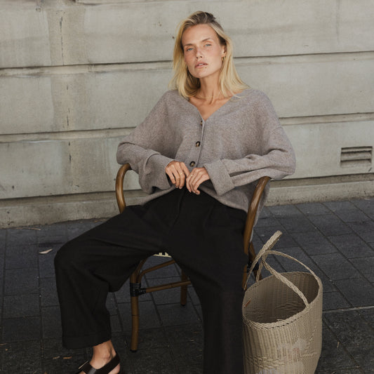 A woman seated wearing black pleated pants with a cuff and double pleats, paired with a grey knit sweater. She is accessorized with a belt and is seated next to a woven basket.
