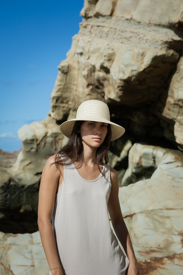 Woman wearing a white dress and straw hat standing in front of rocky cliffs.