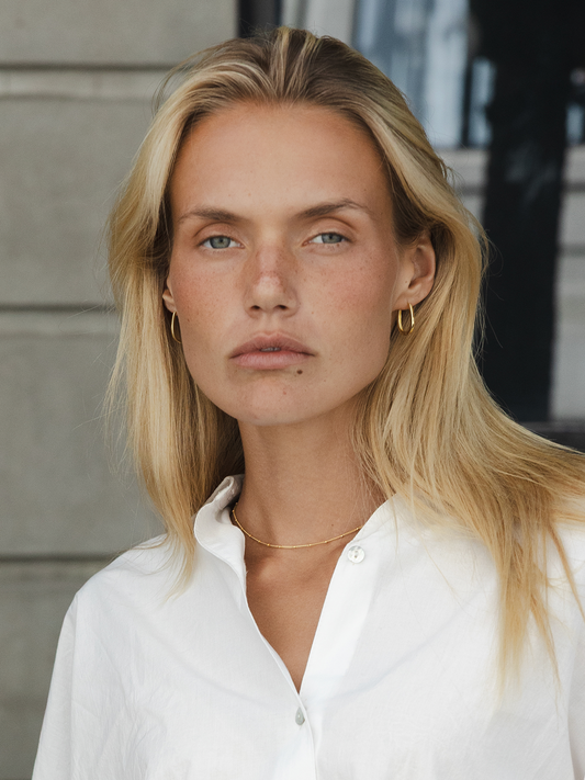 Woman with blonde hair wearing a white shirt, satellite necklace & so sleek hoops against a neutral background