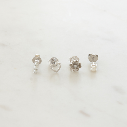 A set of four silver stud earrings with different designs including a heart, a pearl, a floral pattern, and a barbell style on a marble surface.