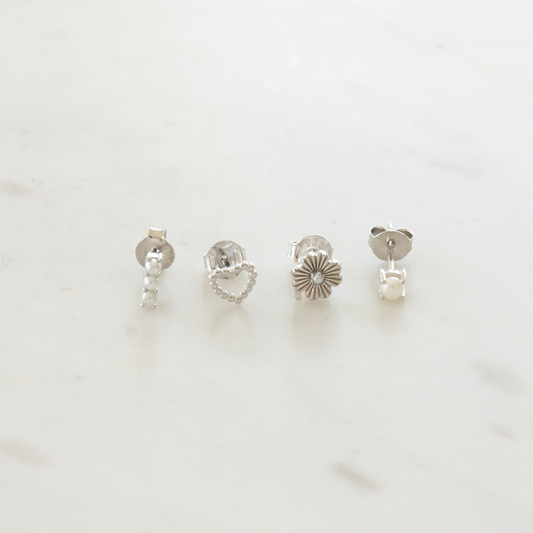 A set of four silver stud earrings with different designs including a heart, a pearl, a floral pattern, and a barbell style on a marble surface.